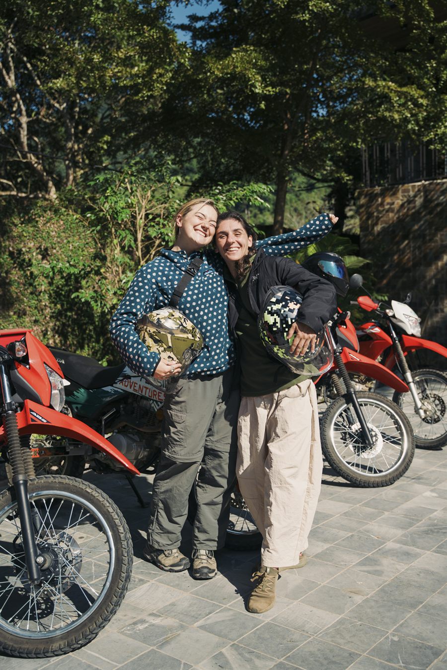 Two riders in gear with their helmets, arms round each other beside the bikes