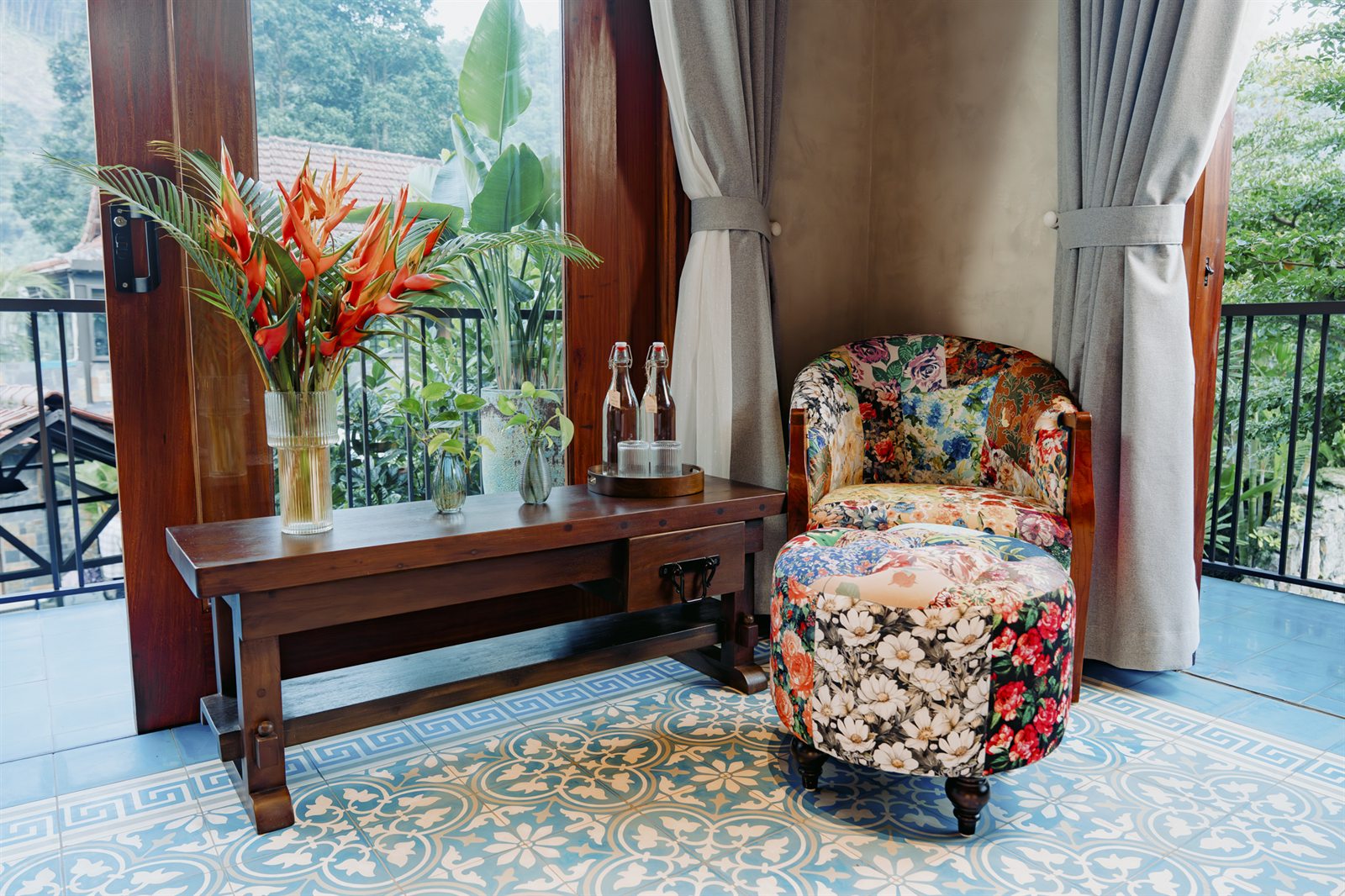 A table of flowers and a floral armchair on a tiled balcony, open to the greenery