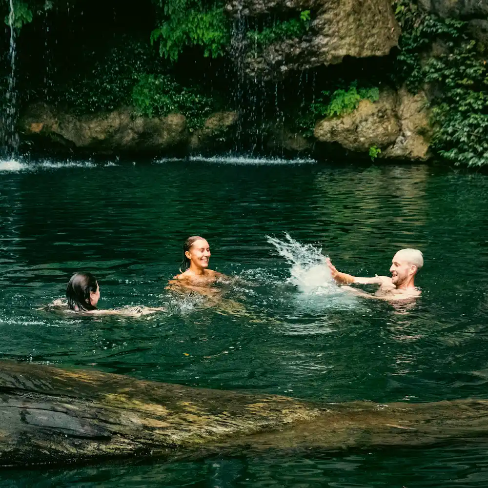 Three riders swimming in a green pool under a waterfall