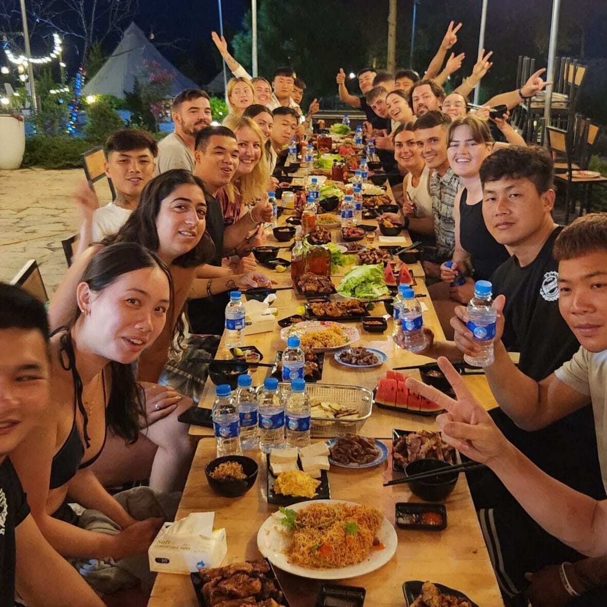 A long table of riders and guides at dinner, hands in the air